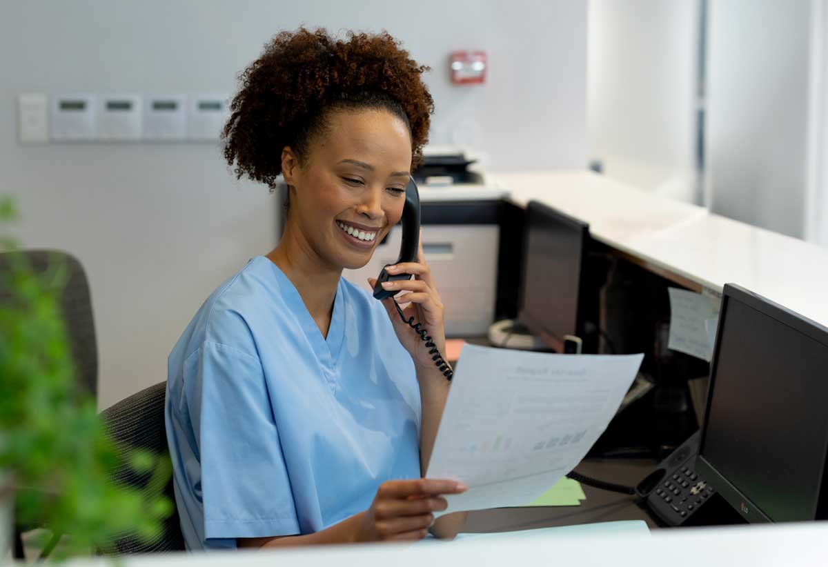A smiling healthcare professional in blue scrubs sits at a desk, talking on the phone while holding a sheet of paper, with computer monitors and office equipment in the background.