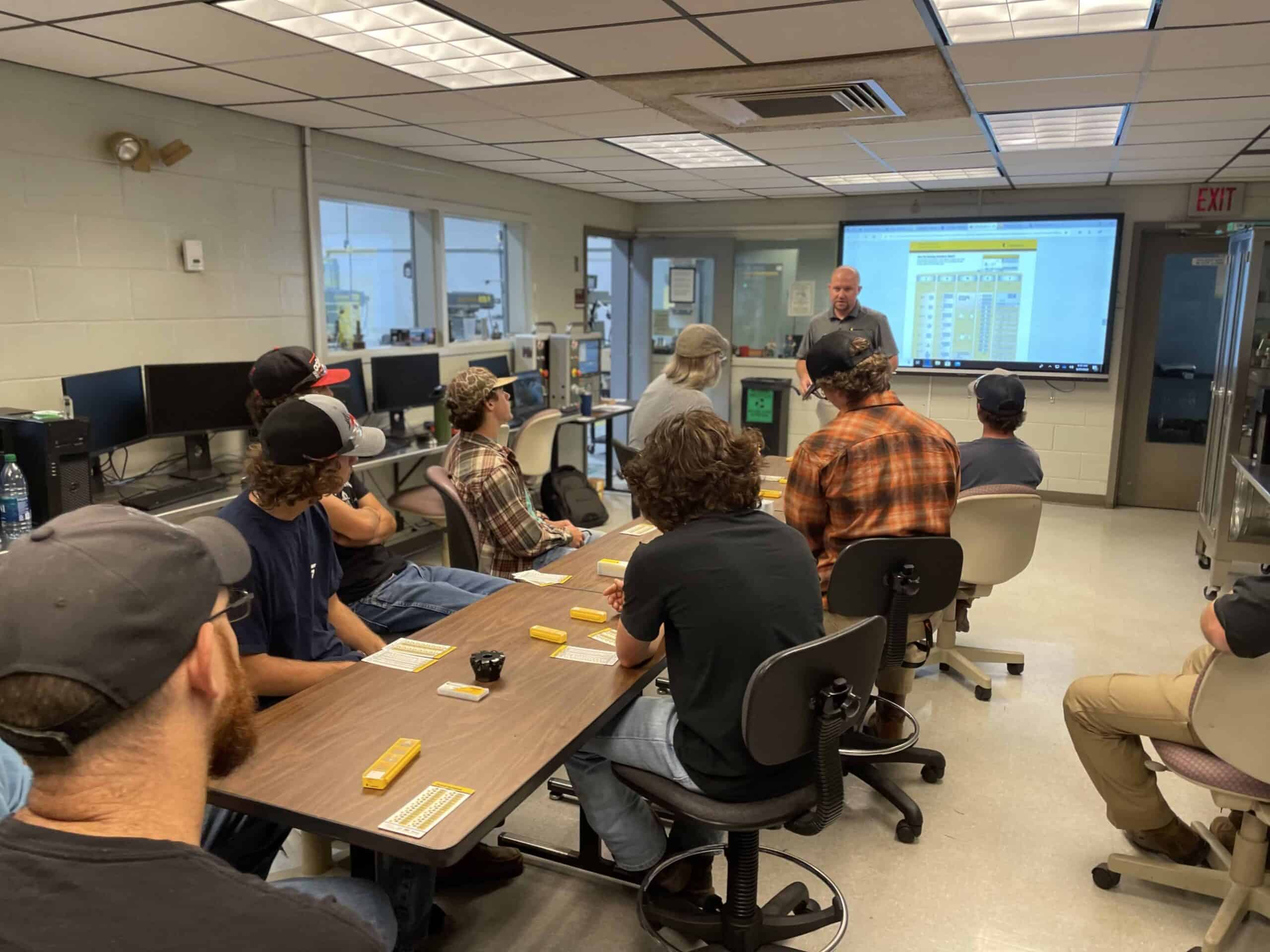 A group of people seated at tables in a classroom listen to an instructor who stands near a screen displaying a yellow diagram. The room has computers, glass windows, and various classroom equipment.