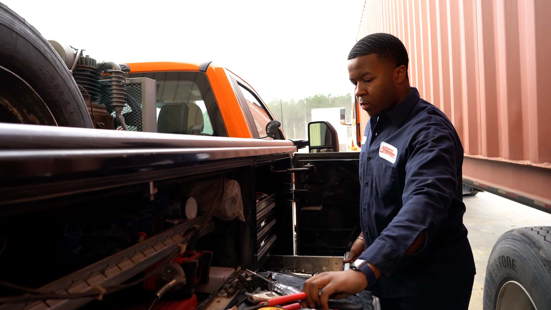 A man in a navy work shirt stands beside an orange truck, using tools from the open side compartment. He appears focused on his task, with shipping containers visible in the background.