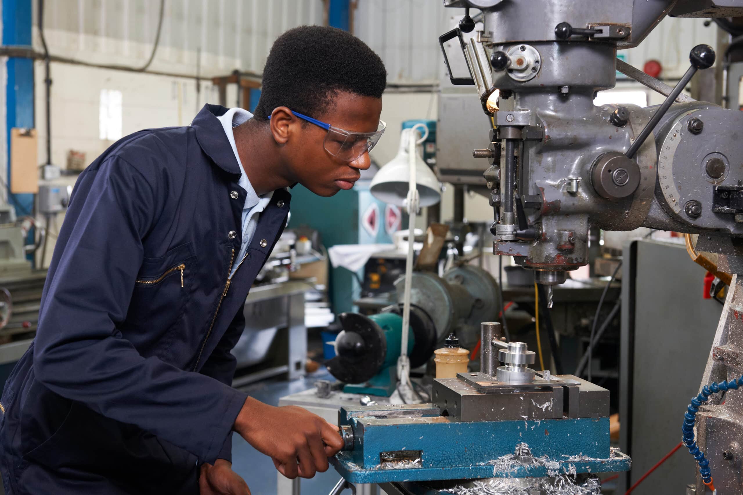 A young man wearing safety glasses and coveralls operates a metal milling machine in an industrial workshop, focusing intently on his work.