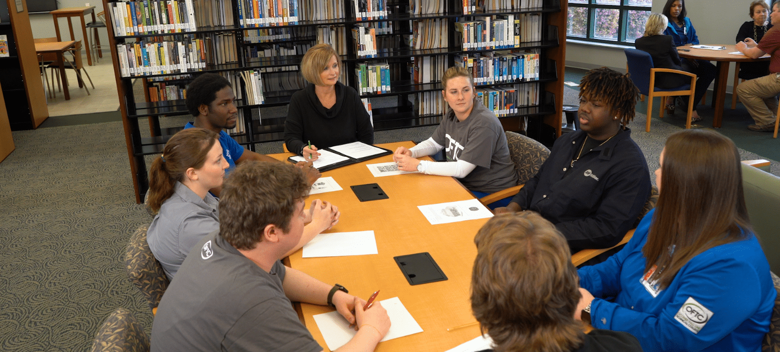 A group of seven people sit around a table in a library, engaged in discussion. Papers and pens are on the table, and bookshelves are in the background. Another small group is visible at a round table by the windows.