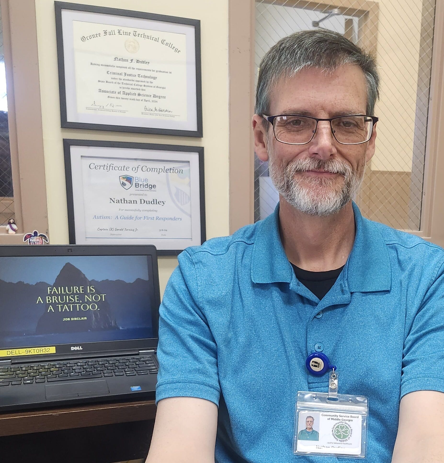 A man with glasses and a beard, wearing a blue polo shirt and ID badge, sits in an office. Behind him are framed certificates on the wall and a laptop with a quote on the screen that reads, Failure is a bruise, not a tattoo.