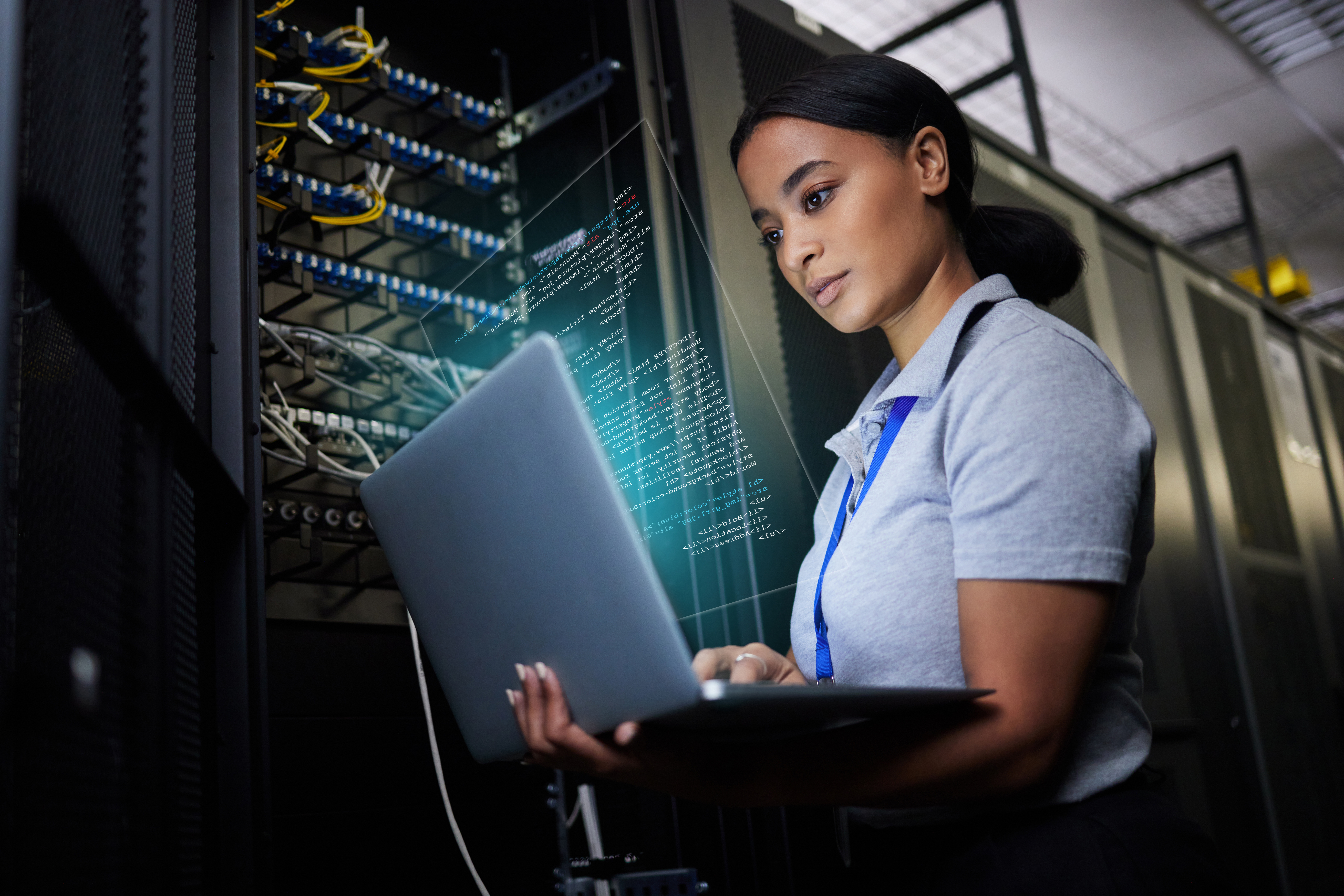 A woman stands in a server room holding a laptop, focused on coding or monitoring data. Digital code is superimposed, suggesting advanced technology or cybersecurity work. Server racks are visible in the background.