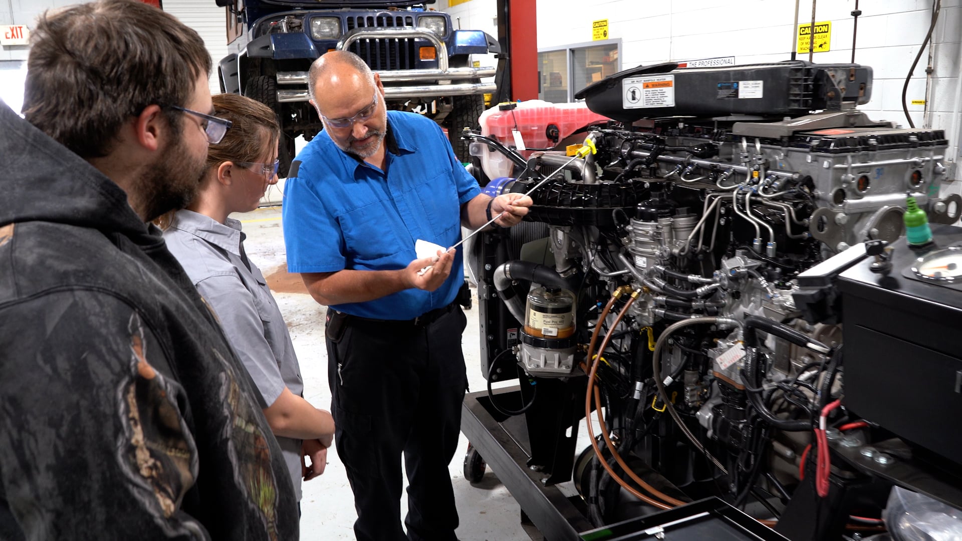 A man in a blue shirt explains engine parts to two students in a workshop, with an exposed vehicle engine and cars visible in the background.