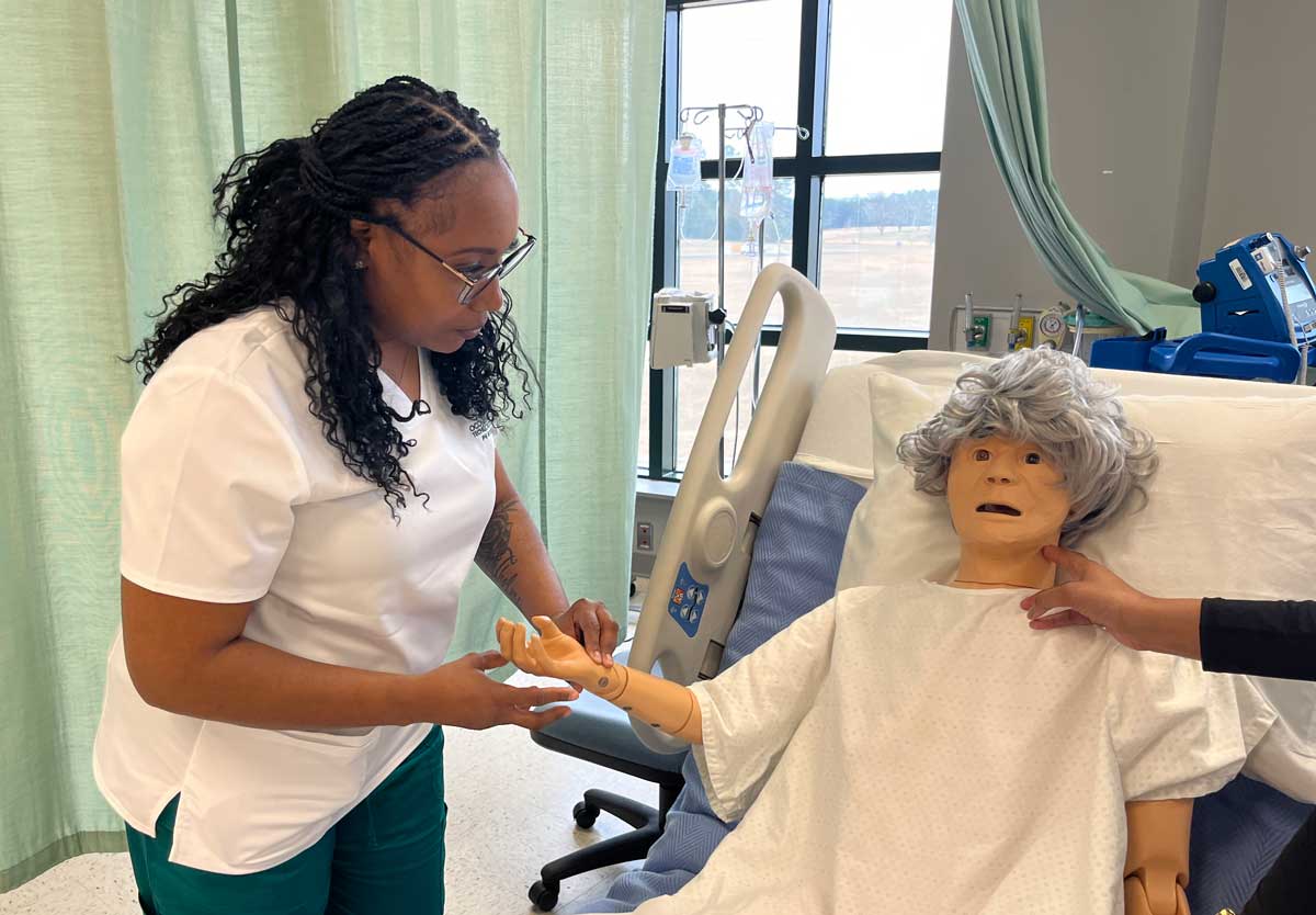 A healthcare worker practices a medical procedure on a life-sized medical mannequin lying in a hospital bed while another person assists by holding the mannequin's arm.