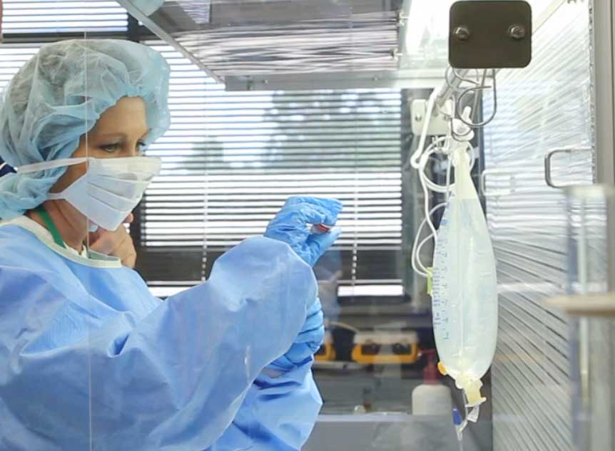 A healthcare worker in protective clothing, mask, and gloves prepares an intravenous (IV) solution inside a sterile laboratory or pharmacy environment.