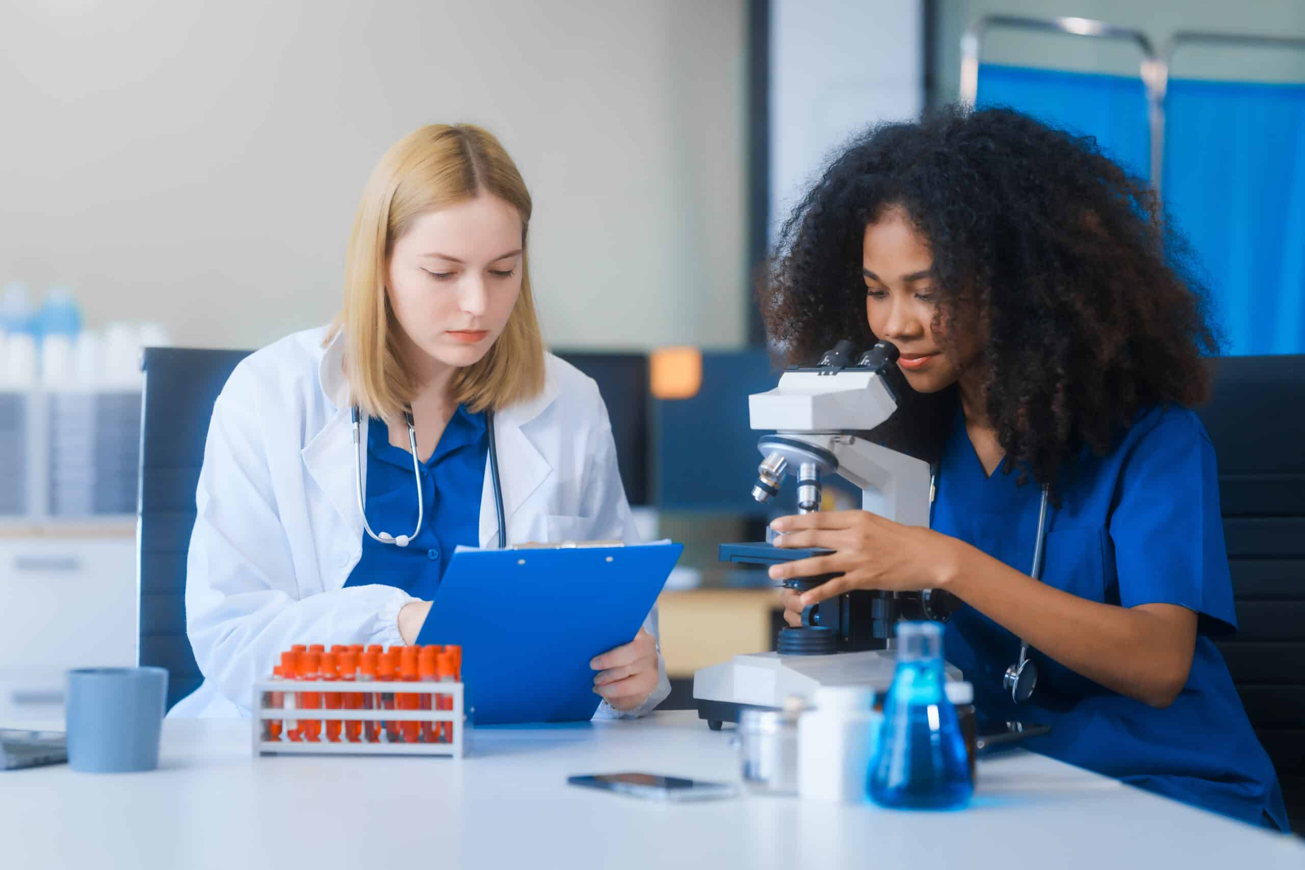 Two female scientists in lab coats work together, one writing on a clipboard, the other using a microscope with test tubes nearby.