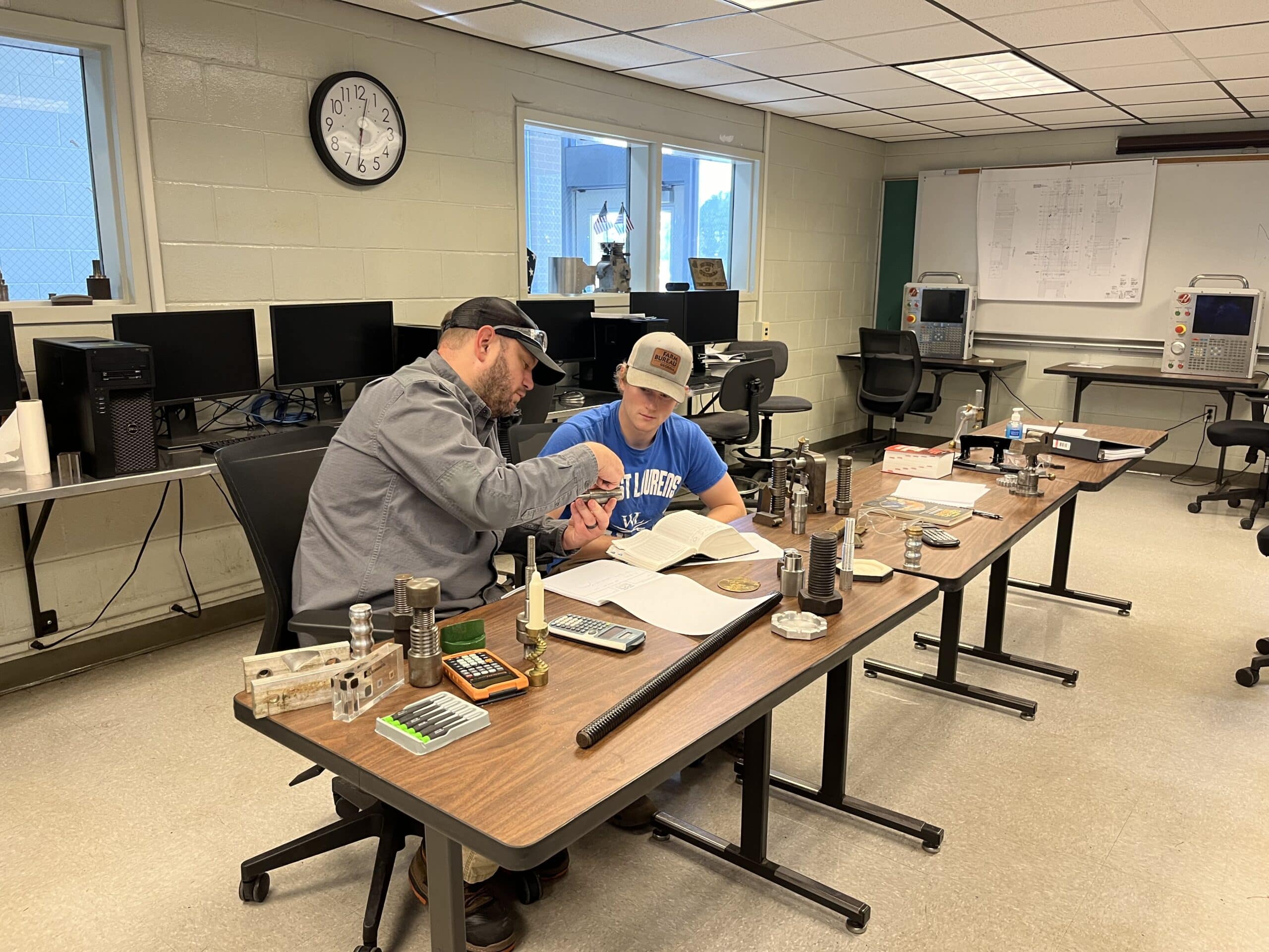 Two people sit at a long table covered with scientific equipment, books, and calculators in a classroom or lab setting. One person points at a notebook while explaining something to the other. Computers and machines are in the background.