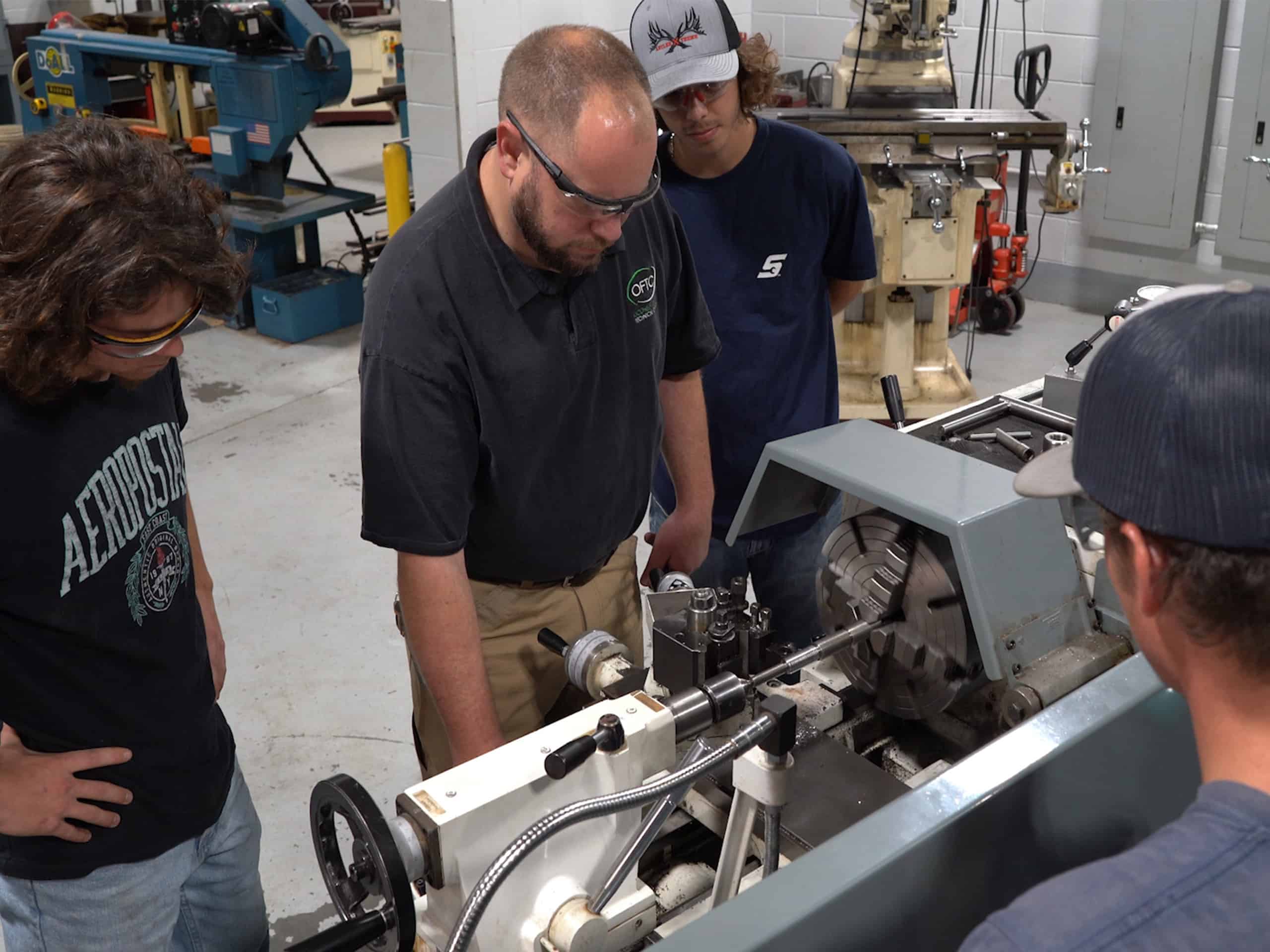 Four people wearing safety glasses stand around and observe a man operating a lathe machine in a workshop with industrial equipment in the background.