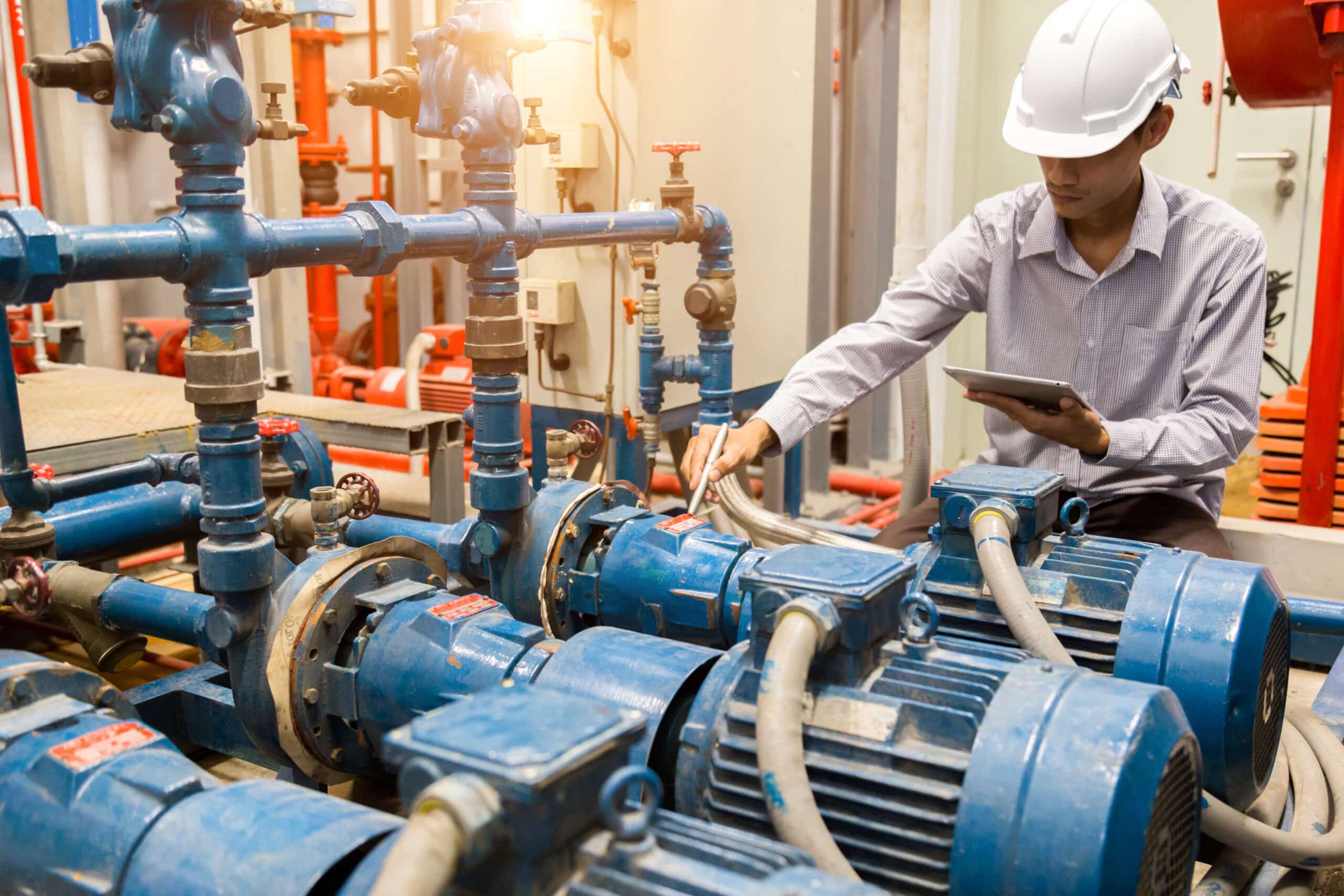 A person wearing a white hard hat uses a tablet while inspecting blue industrial machinery and pipes in a mechanical room.