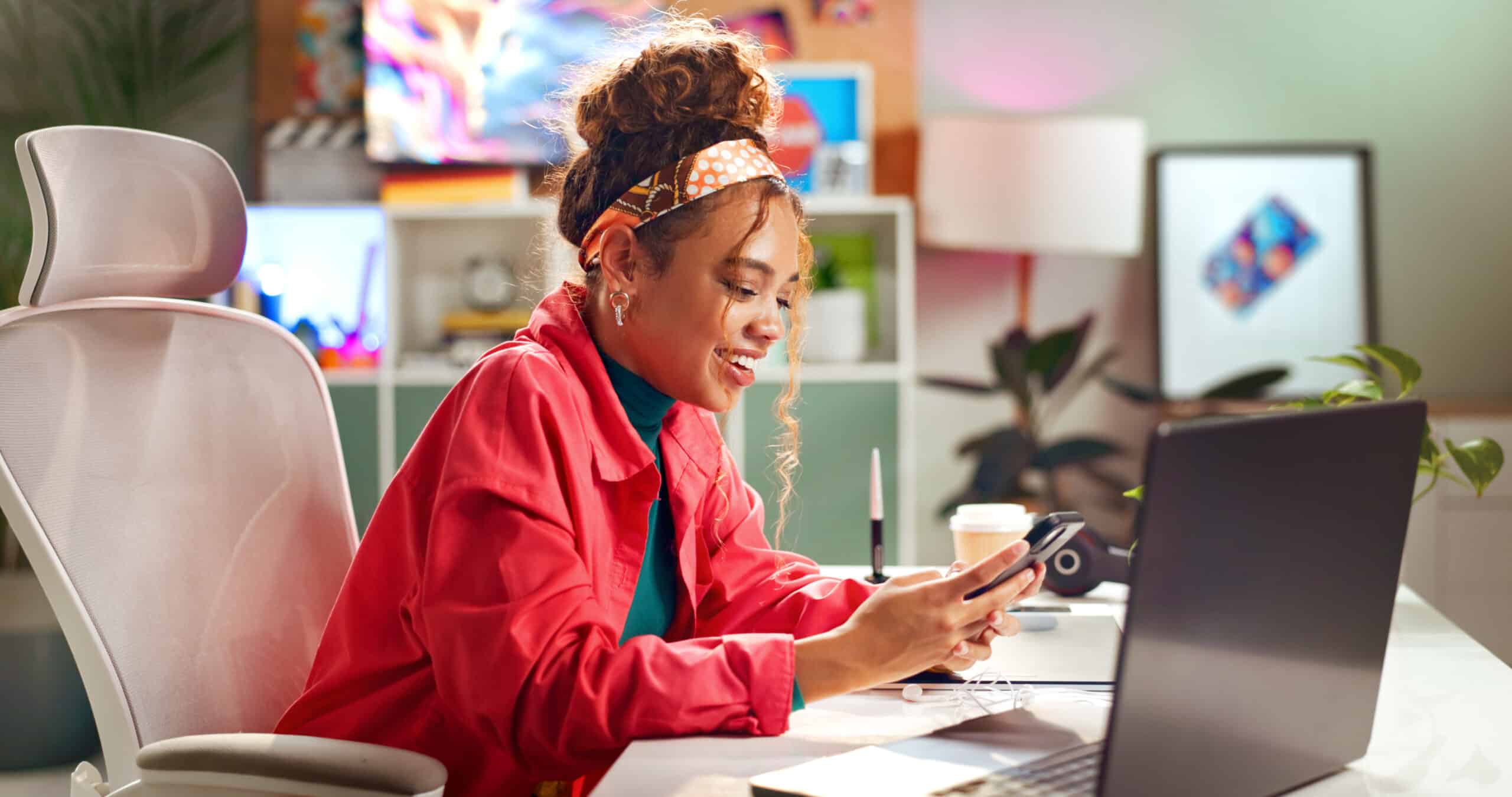 A woman with curly hair in a headband and a red jacket smiles while looking at her phone at a desk with a laptop, notebook, and coffee cup in a brightly decorated office space.