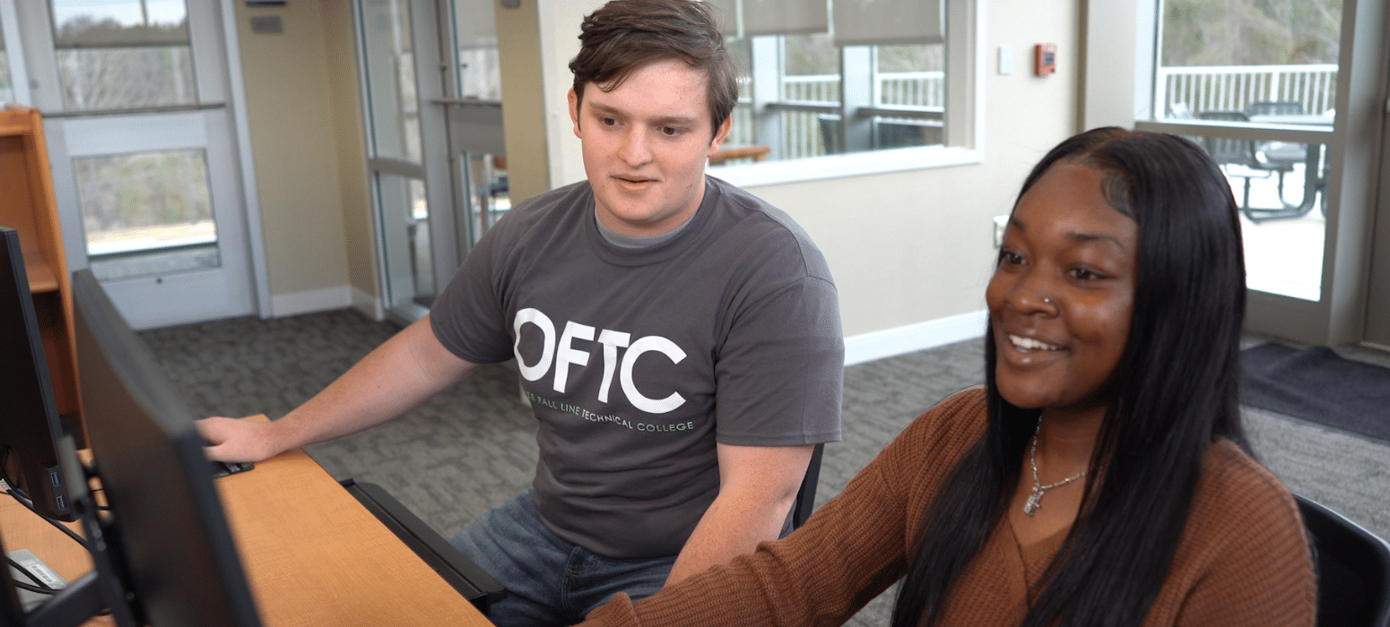 Two students sit side by side at computer desks in a bright room, working together and smiling as they look at the monitors. One wears an OFTC t-shirt and the other wears a brown sweater.