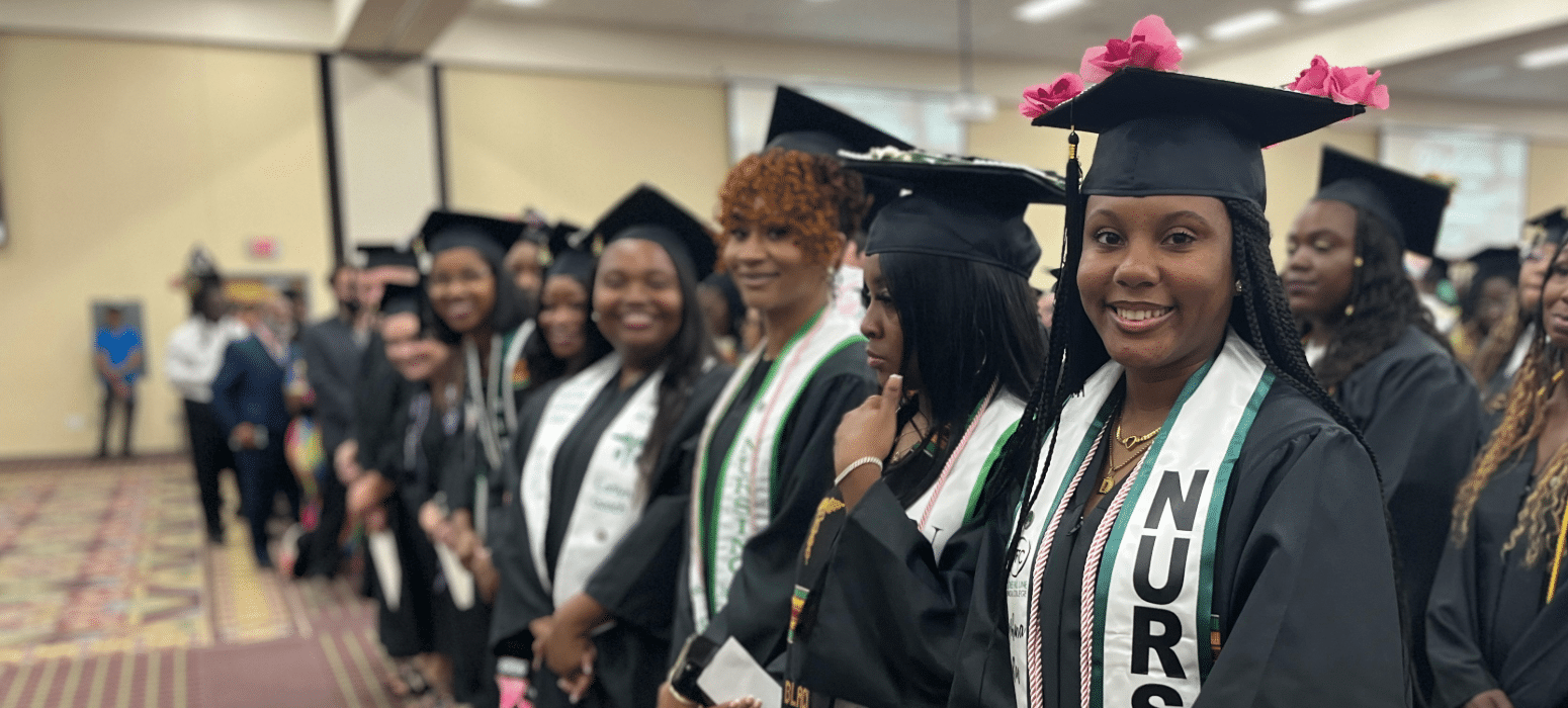 A group of graduates in caps and gowns stand in a row indoors. One woman in front smiles at the camera, wearing a NURSE stole and pink flowers on her cap. Others behind her are also smiling and holding diplomas.
