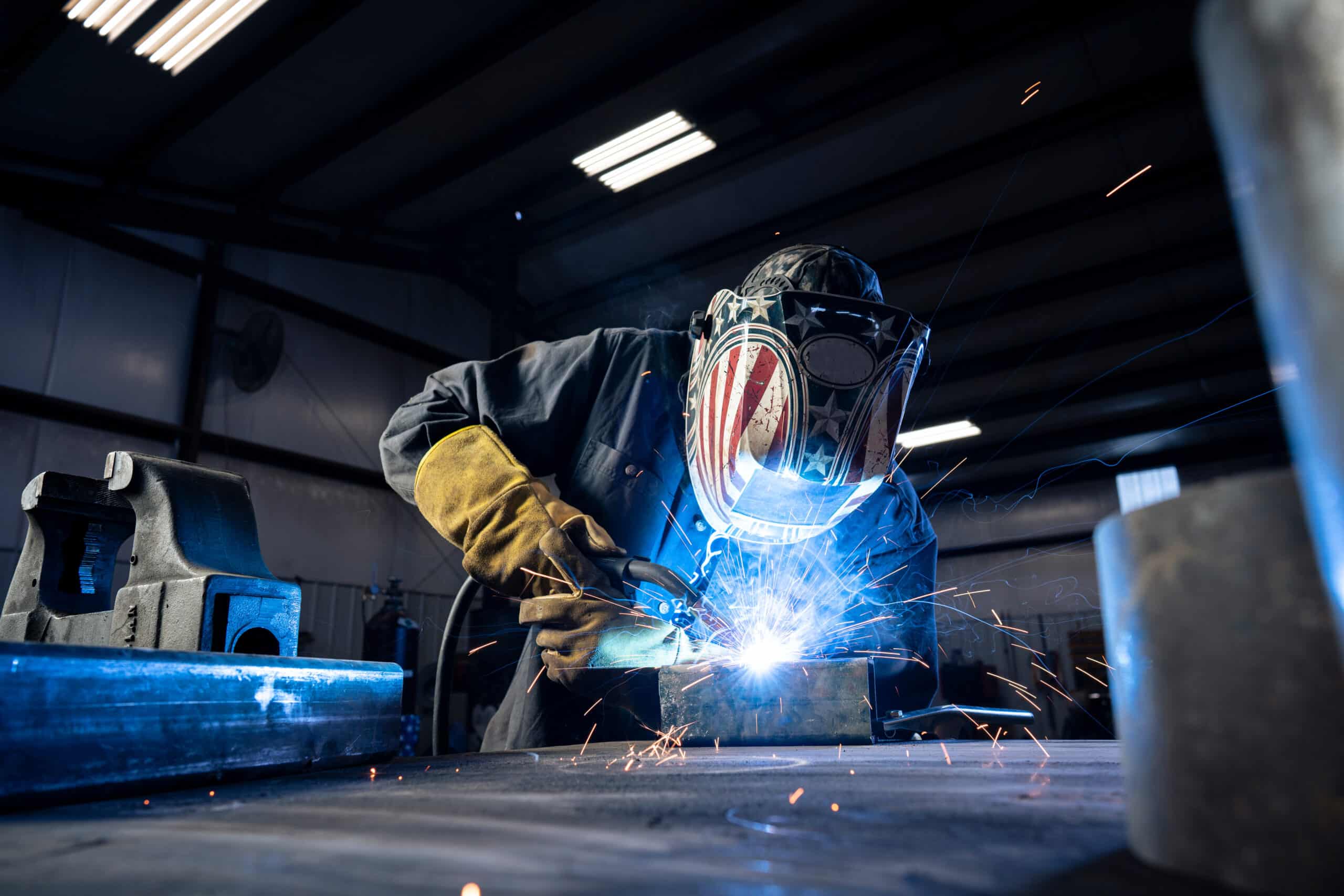 A welder wearing protective gear and a mask with an American flag design is welding metal in a workshop, with bright sparks and blue light illuminating the workspace.