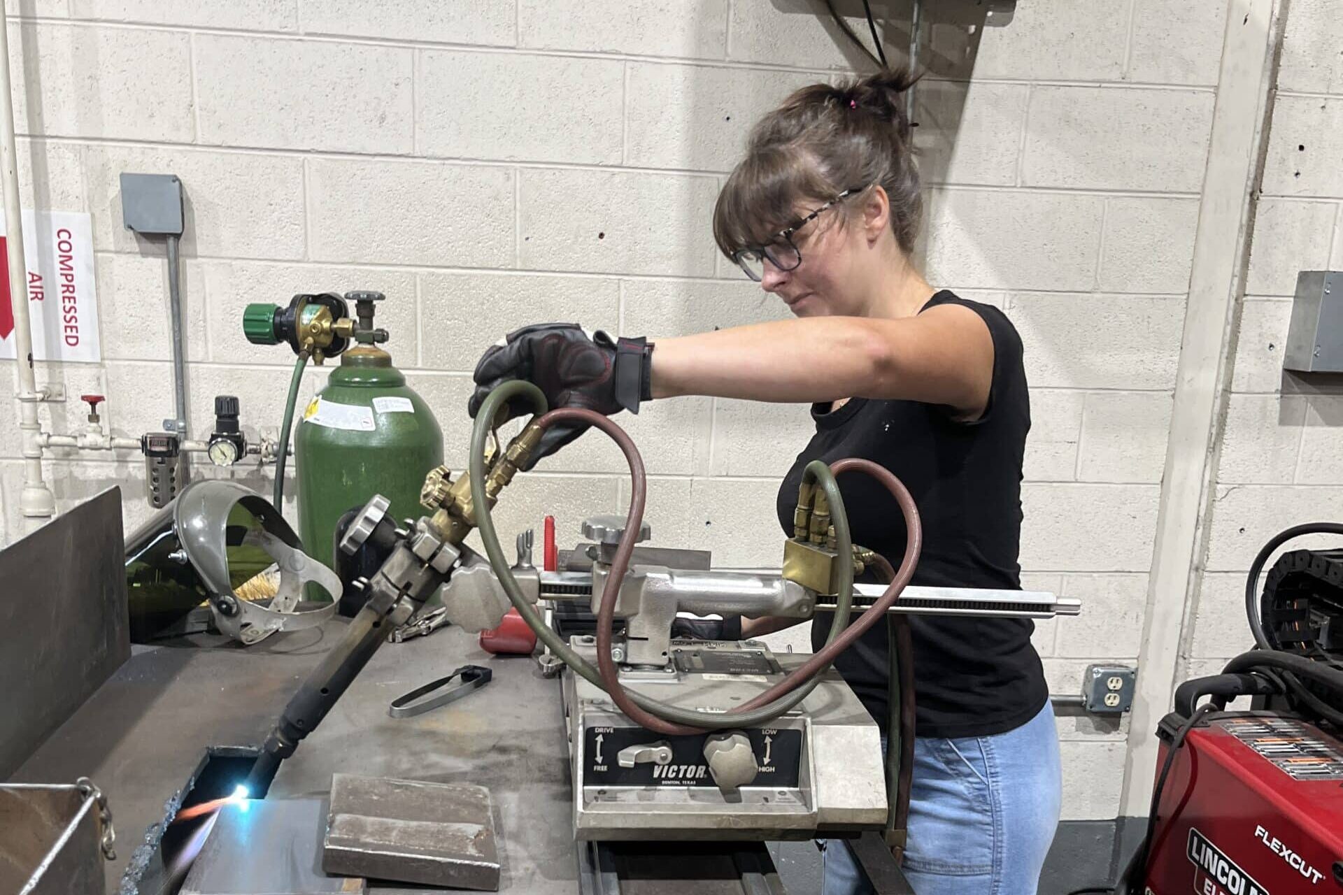 A woman wearing safety glasses and gloves uses a gas torch to cut metal in a workshop, with welding equipment and metal pieces around her.