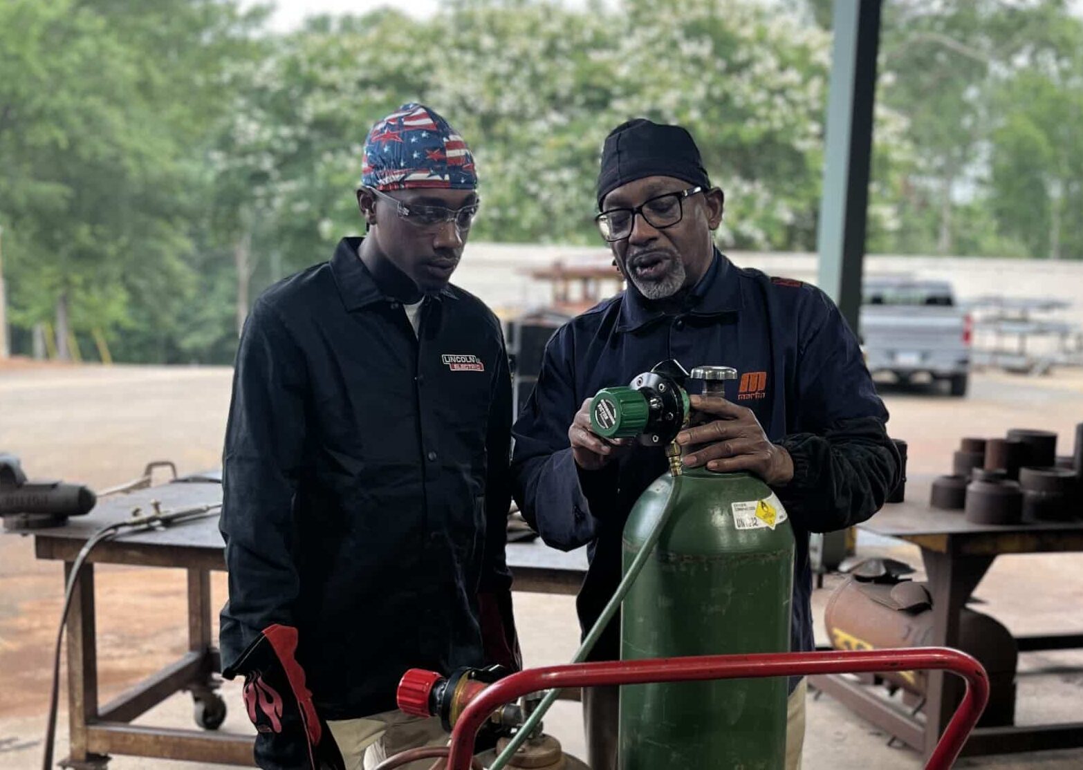 Two men wearing protective gear stand outdoors by a green gas cylinder on a red cart. One man adjusts the valve while the other observes. Industrial equipment and trees are visible in the background.