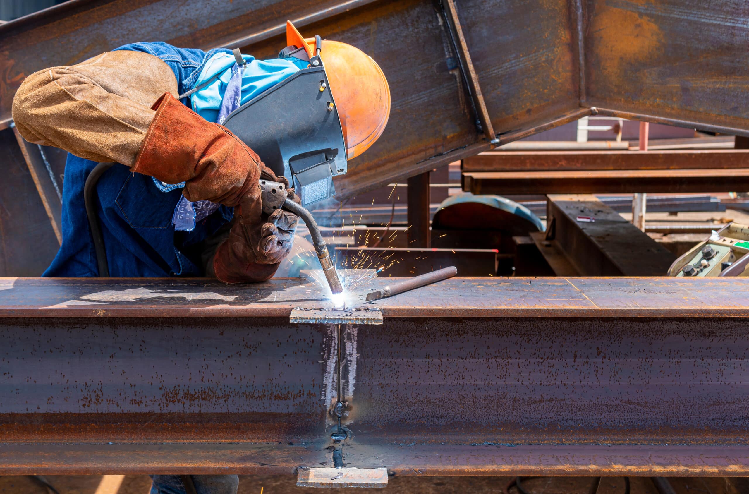 A construction worker wearing protective gear welds steel beams together, with bright sparks flying from the welding torch. Flux Cored Arc Welder