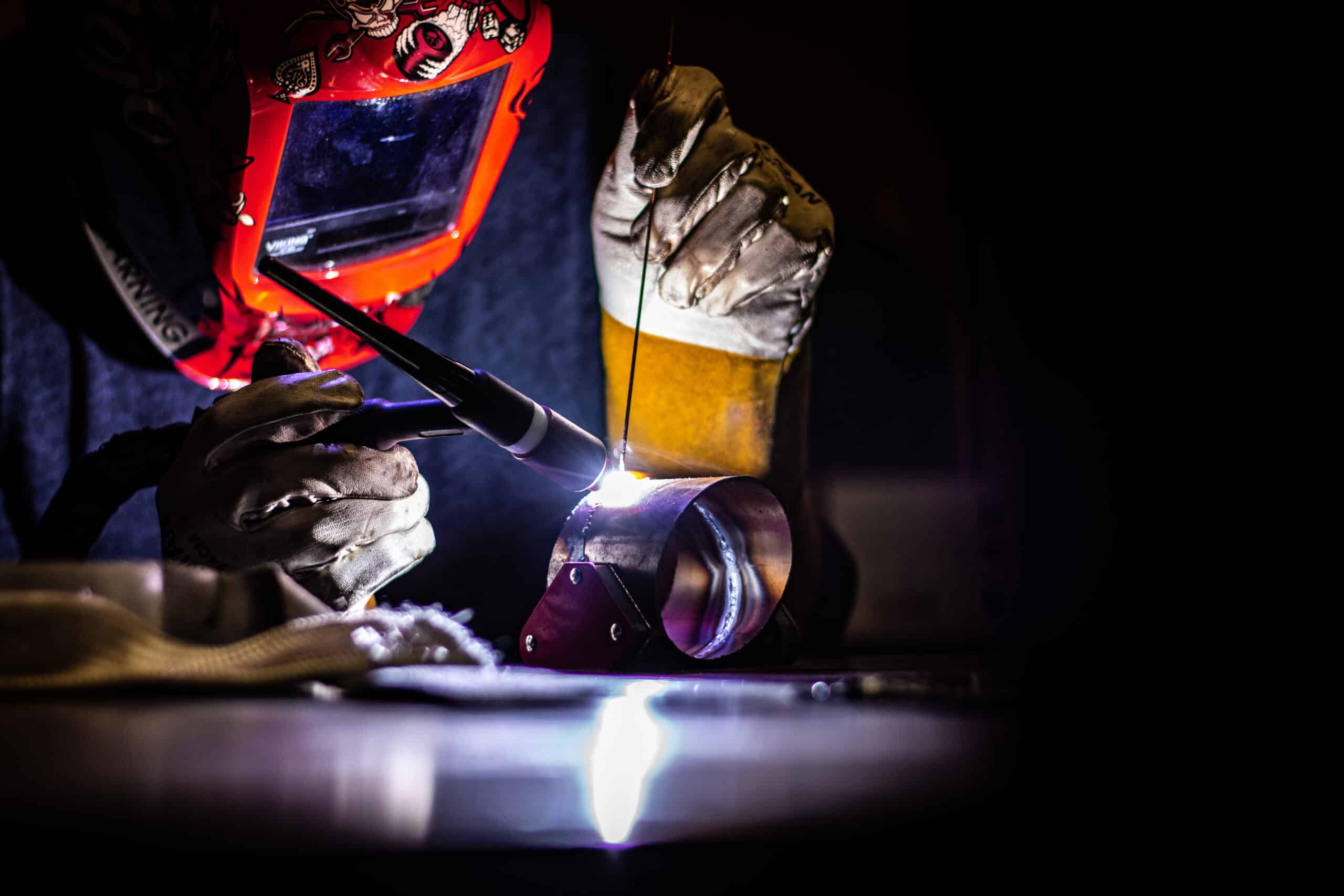 A person wearing a red welding helmet and protective gloves is welding metal, producing bright sparks and a glowing light against a dark background.