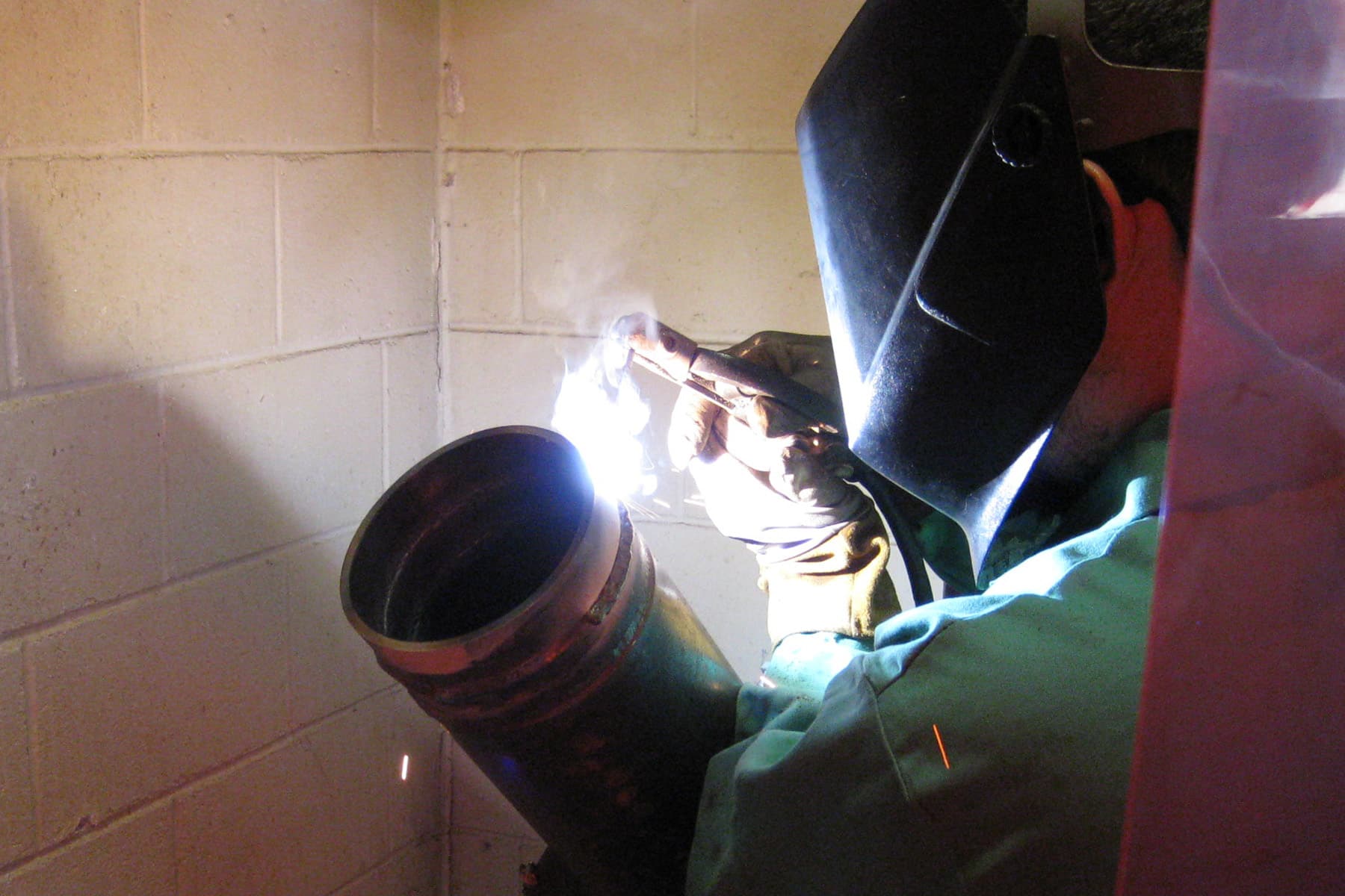 A person wearing protective gear and a welding helmet is welding a pipe in front of a white brick wall, with bright sparks and light visible at the welding point.