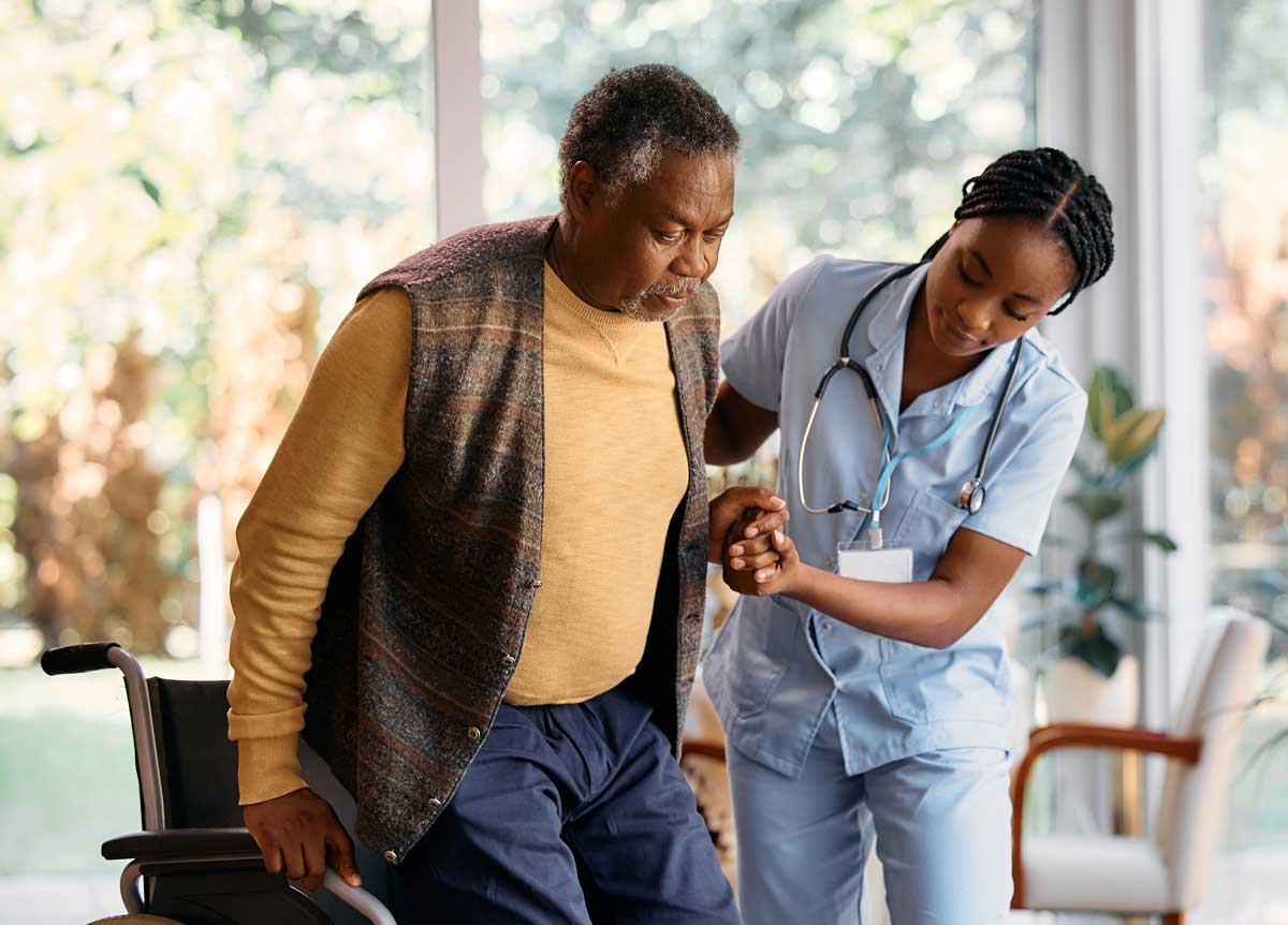 A nurse in scrubs assists an elderly man in standing up from a wheelchair, holding his hand for support. The scene takes place in a bright, indoor setting with large windows in the background.