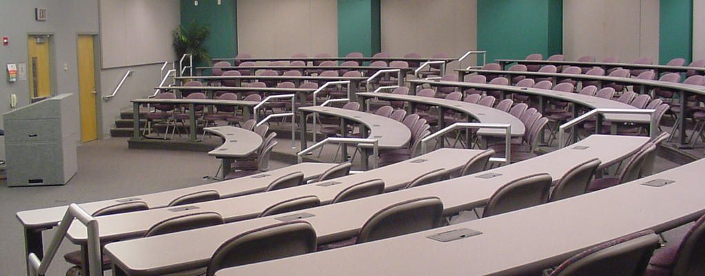 YKK lecture hall with tiered curved desks, metal railings, a front podium, light walls with green accents, and a potted plant.