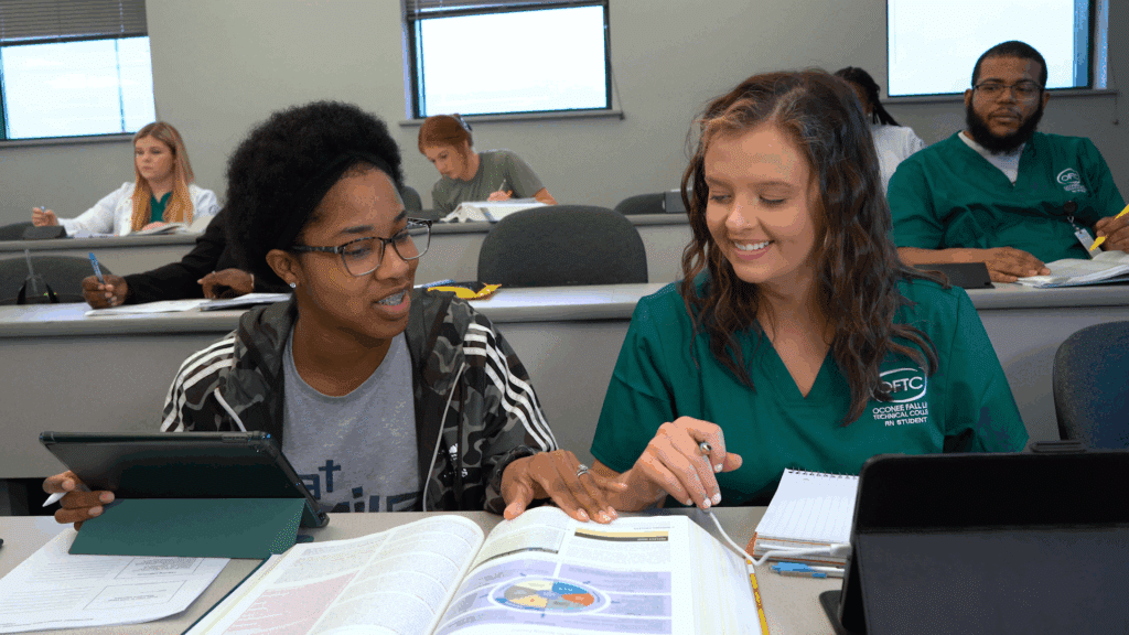 Two students sit at a desk in a classroom with open textbooks and a tablet; one in green smiles, others study in the background.