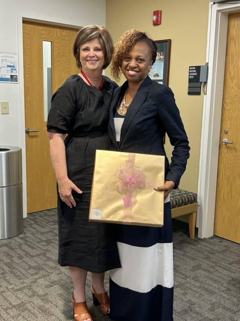Two women smile in an office; one in a black dress and sandals, the other in a blazer and striped dress holds a gift with a pink bow.