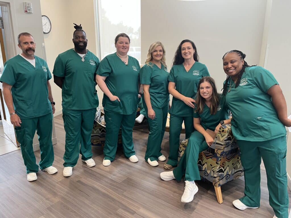 Seven people in green scrubs and white shoes pose together in a bright room, with some standing and two seated on patterned chairs.