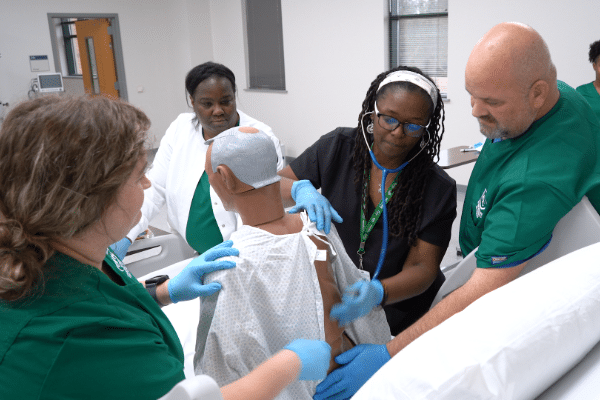 Four medical professionals practice a procedure on a mannequin in a clinical training room; two wear green scrubs, one wears black.