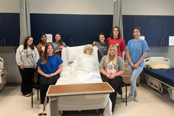 Eight students gather around a hospital bed with a medical mannequin in a classroom, seated and standing; cabinets in background.