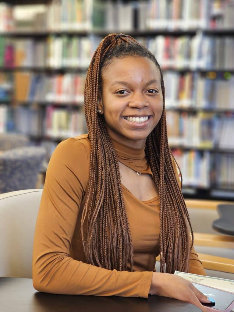 A young woman with long braids, wearing a brown top, smiles as she sits at a table in a library with bookshelves behind her.