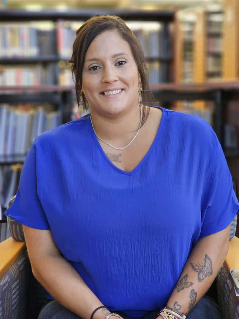 A smiling woman with long brown hair in a bright blue blouse sits in a library, tattoos visible, wearing bracelets and necklaces.