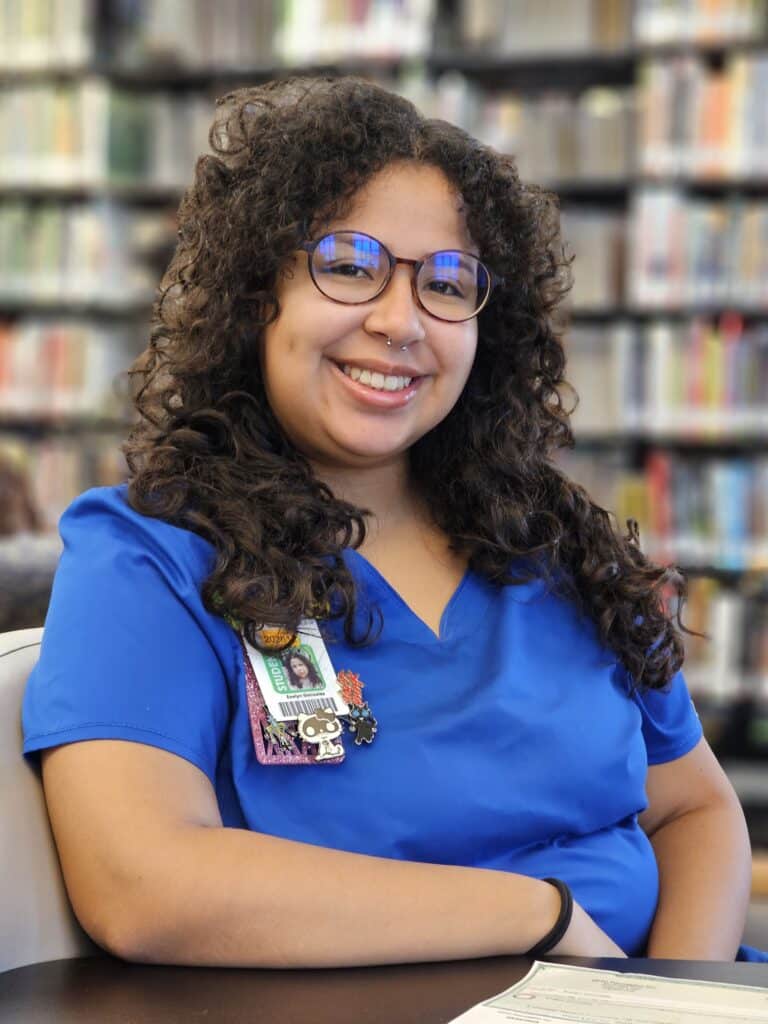 A woman with curly hair, glasses, and a nose ring in blue scrubs and a name badge smiles at a table in a library.