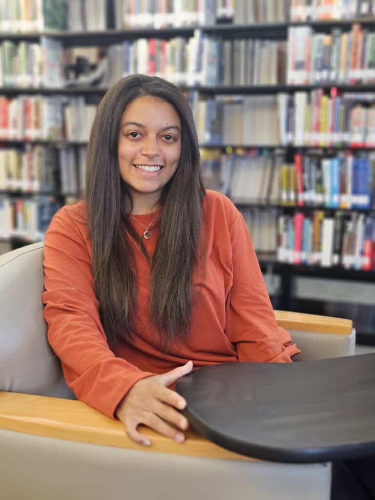 A woman with long straight hair in an orange long-sleeve shirt smiles while sitting in a library chair, bookshelves behind her.