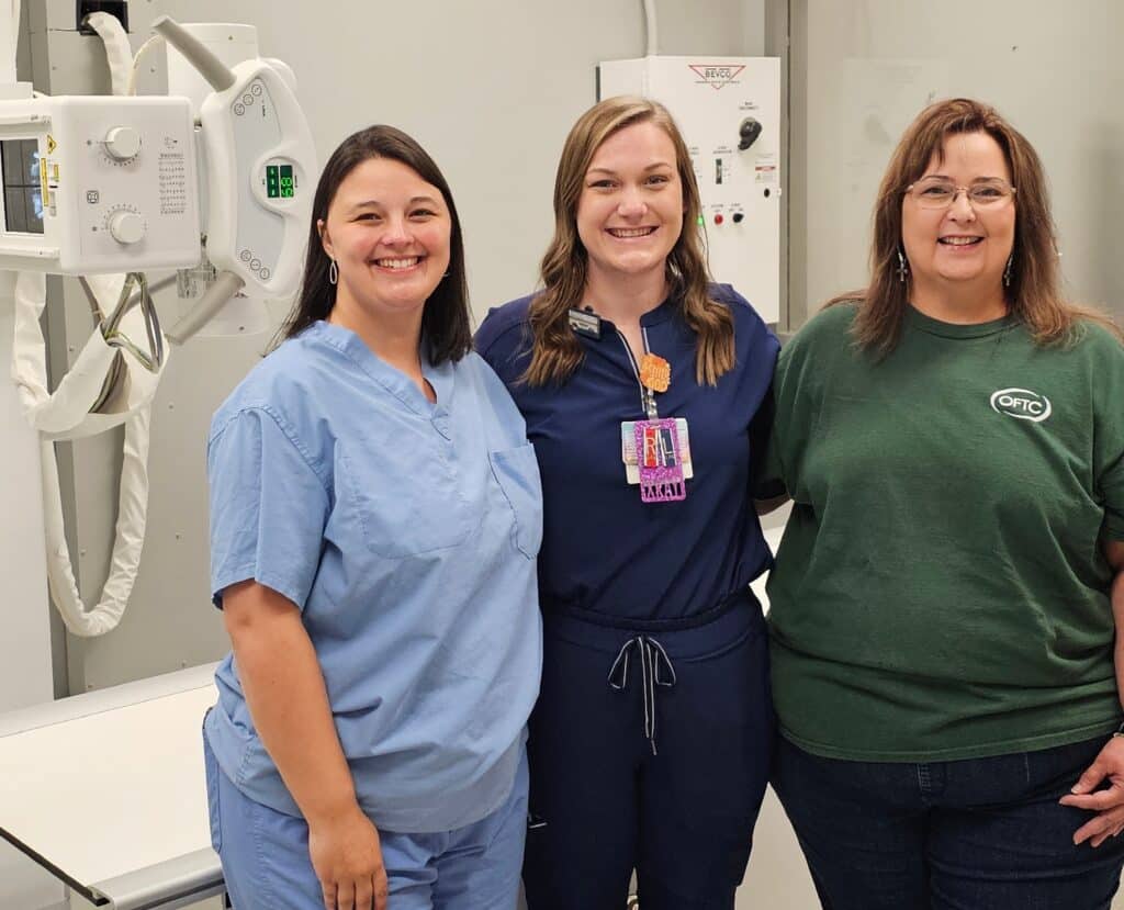 Three women smile together in a medical setting; two wear blue scrubs, one wears a green t-shirt, with equipment behind them.