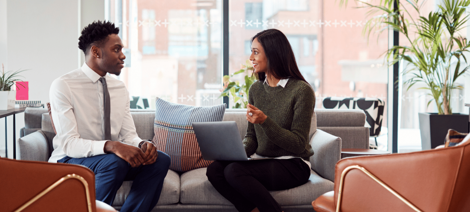 Two people sit on a sofa in a modern office, talking. One gestures with a laptop; the other listens. Windows and plants behind them.