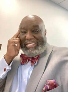 Speaker Demetrius Smith - A cheerful gray-bearded man in a grey suit, colorful bow tie, and matching pocket square poses indoors, hand to his face.