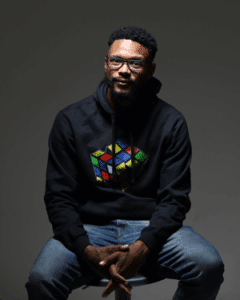 Speaker Dr Derrick-Standifer - A man with short curly hair and glasses sits on a stool, wearing a black hoodie with Rubik’s Cube graphic and blue jeans.