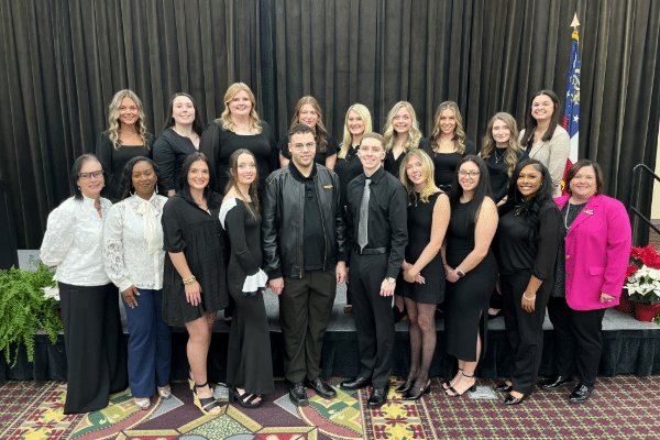 A group of 17 people, mostly women in black with a few in white or pink, pose indoors before black curtains and an American flag.