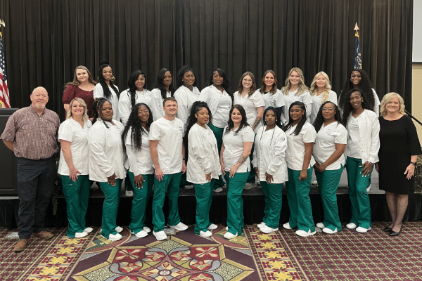 A group of mostly women in white tops and green pants pose indoors with three in business attire, in front of a black curtain and two US flags.