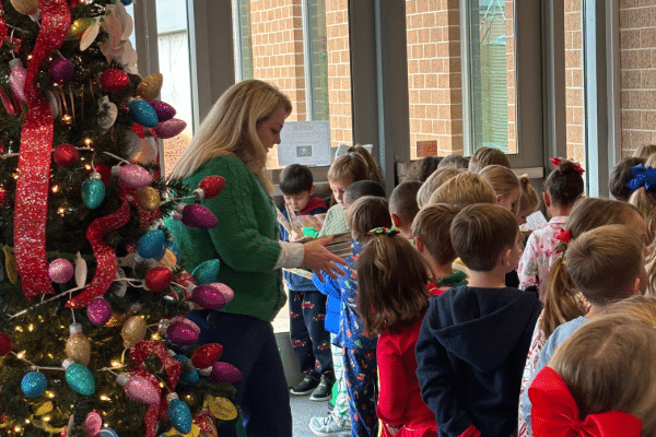 A woman in a green sweater stands by a Christmas tree, leading young children in colorful clothes lined up indoors near big windows.
