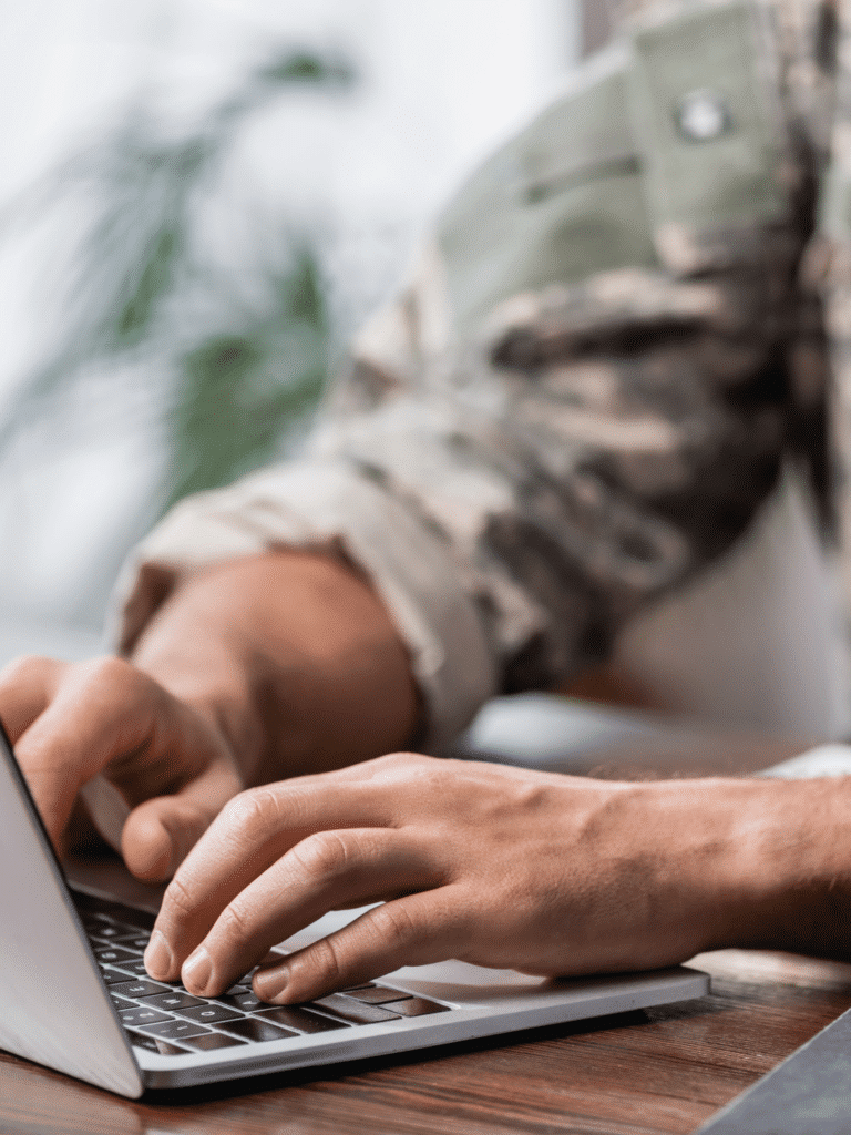 A person wearing a camouflage military uniform uses a laptop at a desk, showing only their hands and forearms.