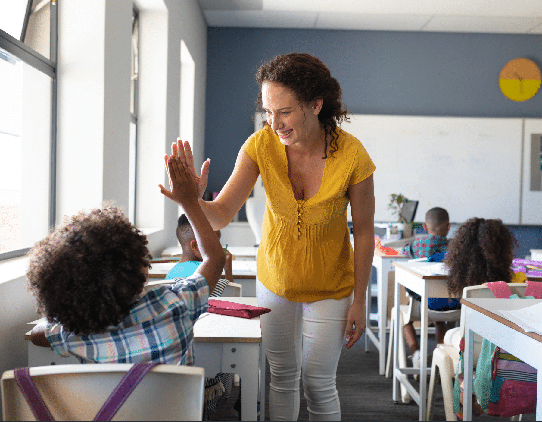 A teacher in a yellow blouse smiles and high fives a student at a desk in a bright classroom with other children in the background.
