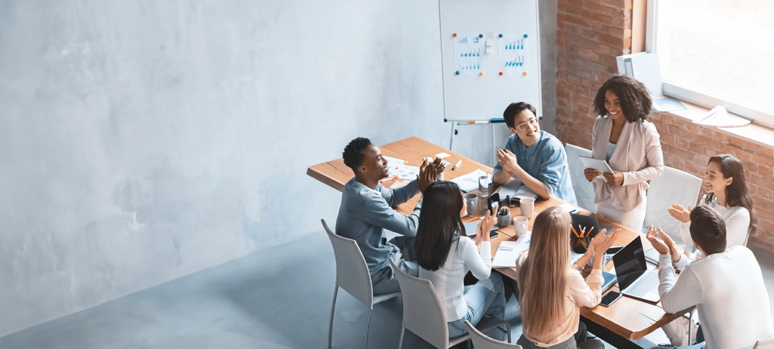 Six people sit around a conference table in a modern office, one standing and speaking while others listen and take notes; graphs on whiteboard.