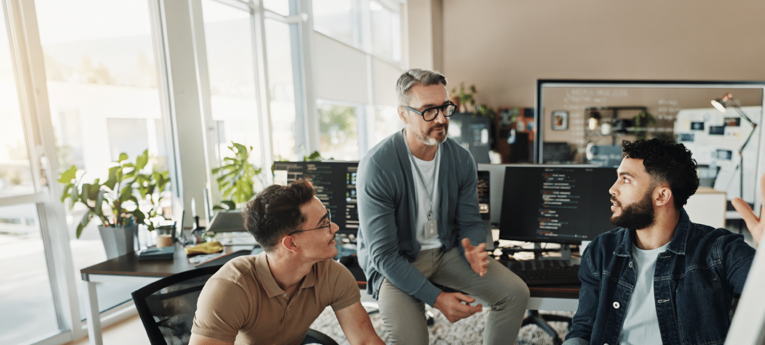 Three men discuss near computer monitors showing code in a sunlit modern office with large windows and visible indoor plants.