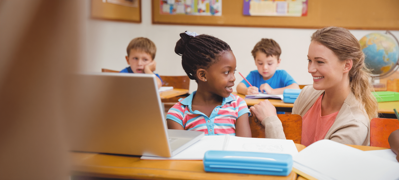 A teacher smiles while talking to a young girl in a classroom, with other students working at their desks and supplies on the desk.
