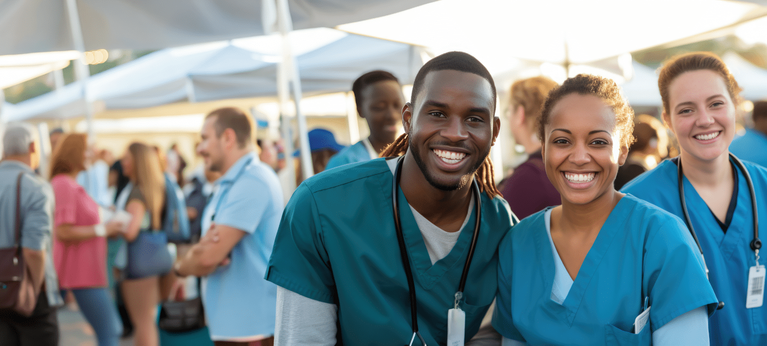 Smiling healthcare workers in scrubs stand together outside at a community event, with tents and people visible in the background.