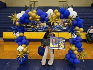A young woman in a gym smiles, holding a diploma, gift bag, and a poster with her graduation photo and Chatham Class of 2026.