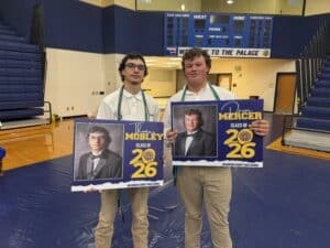 Two high school students in white shirts and cords stand in a gym, each holding a Class of 2026 senior poster with their names.