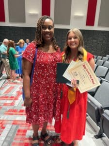 Two women smile at a graduation; one in a red dress with blue bag, the other in gown with gold stole holding certificates. People chat behind.